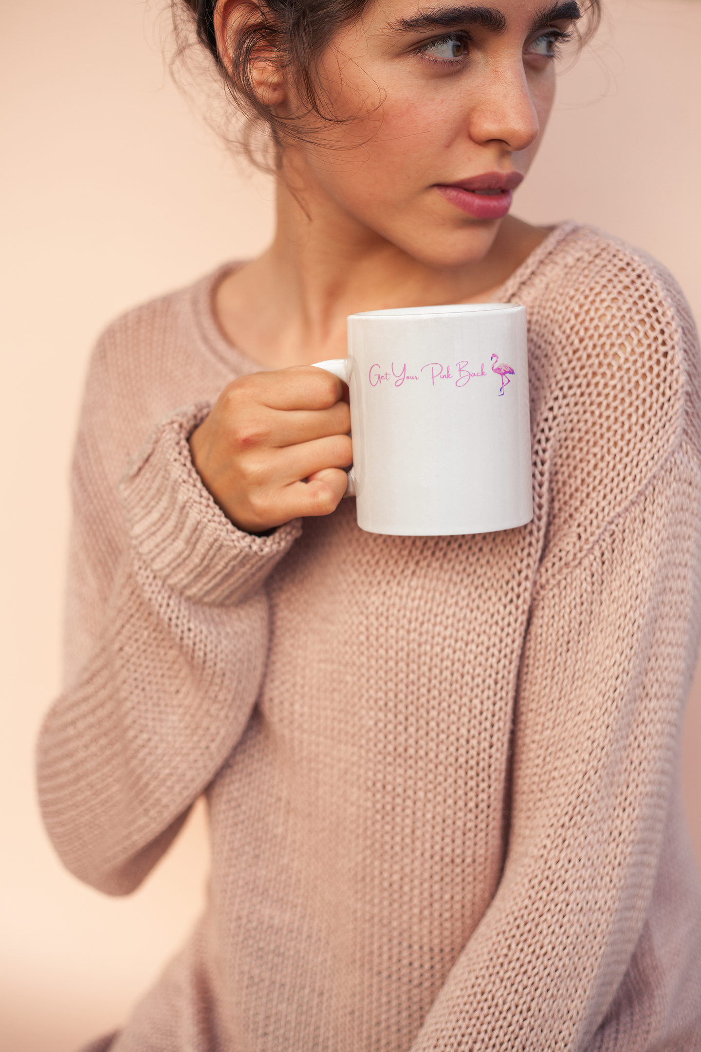 Woman holding a white mug with text, wearing a pink sweater against a beige background