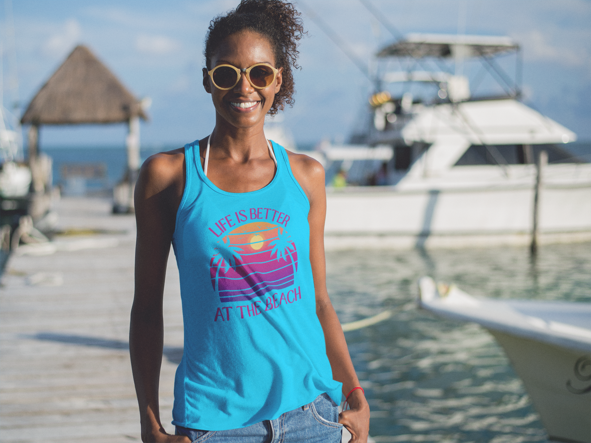Woman wearing a blue tank top with a beach-themed design, standing on a dock with boats and water in the background.